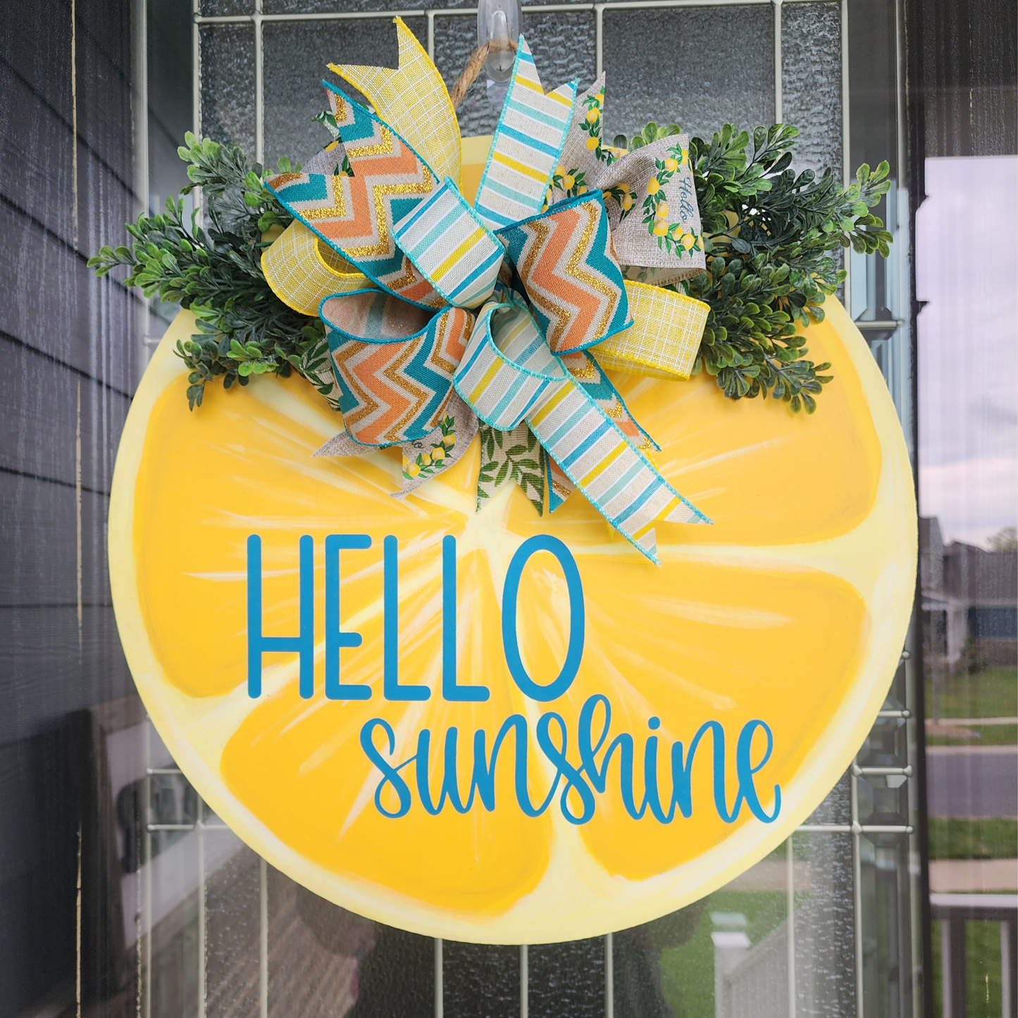 18 inch birch decorative door round painted to look like a lemon slice with the words hello sunshine in teal. Colorful bow and greenery in the top center.